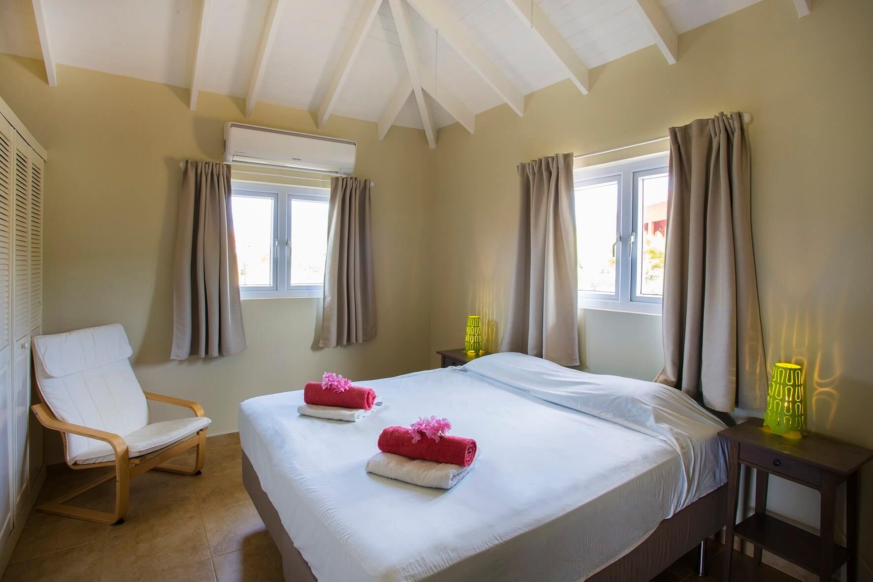 Een slaapkamer met opgemaakt tweepersoonsbed, fauteuil en twee ramen